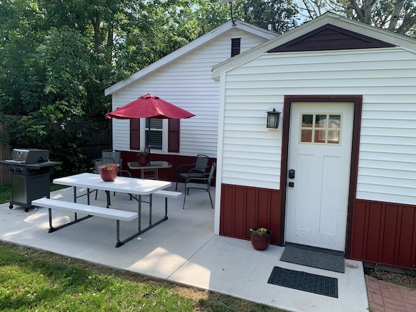 A small, single-story house with a white exterior and brown trim. It features a patio with a picnic table and chairs, a red umbrella, and a grill.