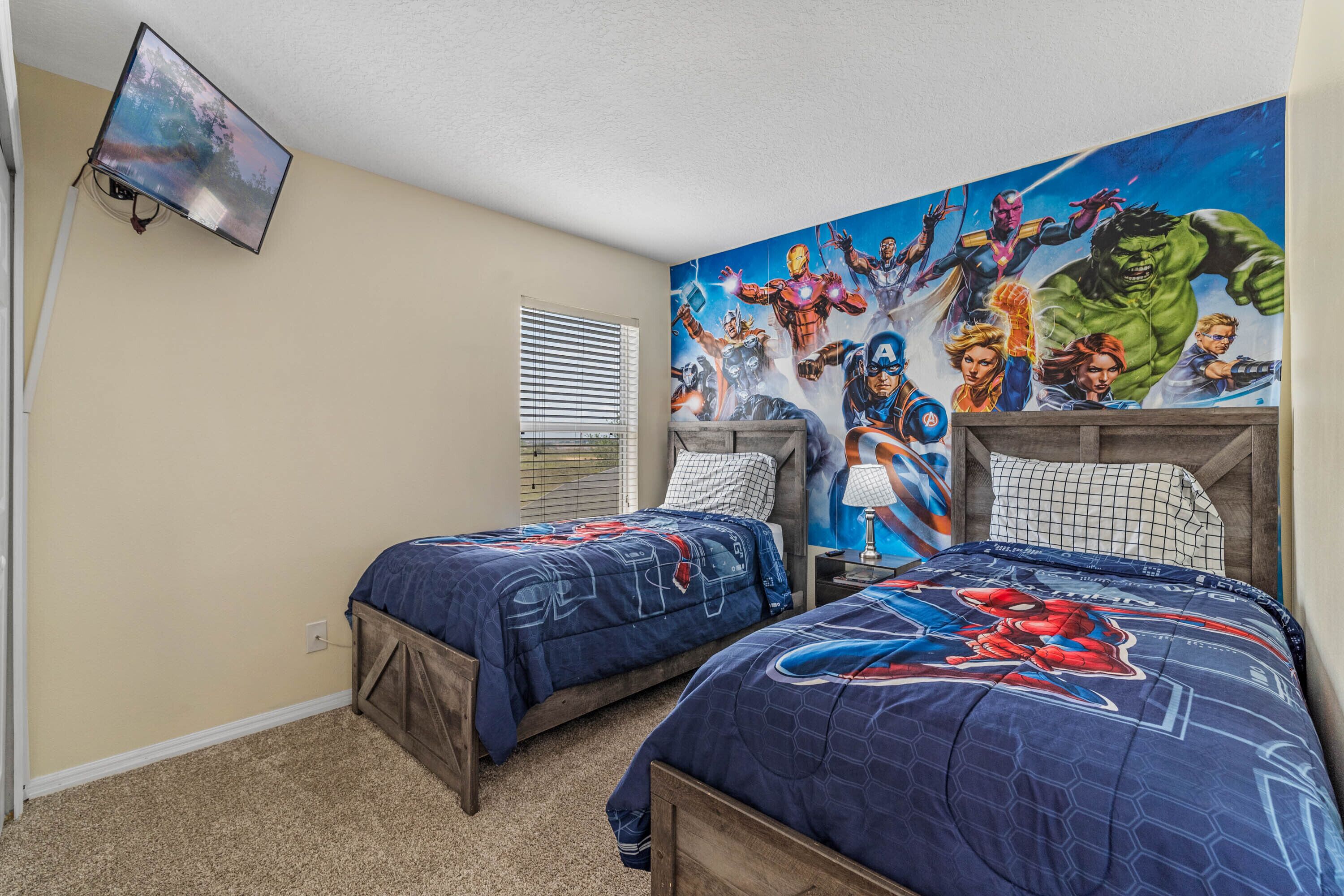 A bedroom with two beds, a mural of superheroes on the wall, a TV mounted on the wall, and a window with blinds.