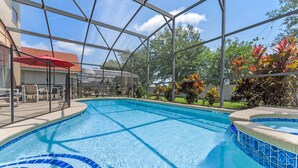En swimmingpool med et glasafskærmet område, omgivet af en terrasse med udendørs møbler og et overdækket areal.
