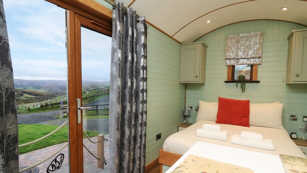 A bedroom with a bed, a painting, a window with curtains, a view of the outdoors, and a wooden door. - Highland View (Rhayader)