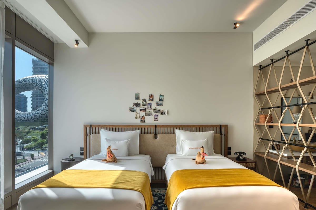 Hotellihuone, jossa on kaksi sänkyä, suuri ikkuna, valokuvilla koristeltu seinä ja koristeellinen hylly.