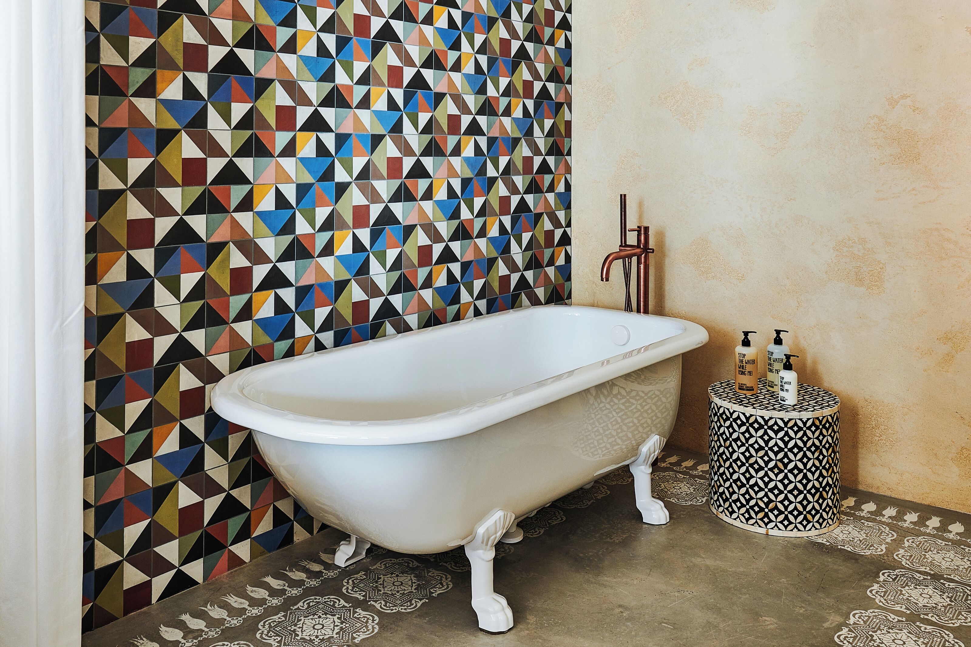 A bathroom with a white clawfoot bathtub, a patterned wall, and a round side table with toiletries.