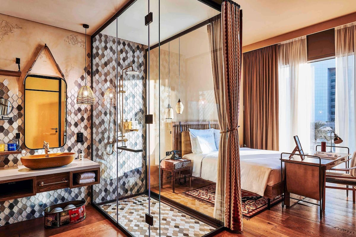 A hotel room with a glass partition, a bed, a desk with a chair, a bedside table, and a large window.
