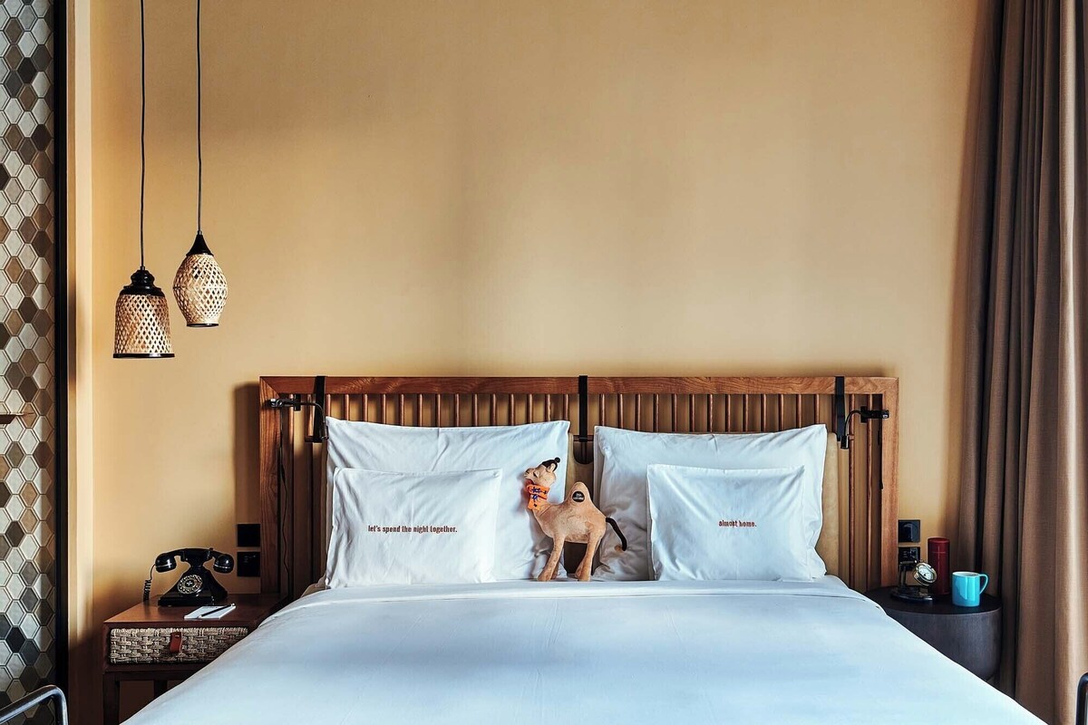 A hotel room with a wooden headboard, two white pillows with text, a small dog figurine, a vintage telephone, and a nightstand with a blue mug.
