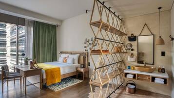 Una habitación de hotel moderna con una cama de madera, un escritorio, un ventanal con cortinas, una estantería geométrica original, un espejo redondo y un lavamanos de madera.