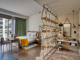 Una habitación de hotel moderna con una cama de madera, un escritorio, un ventanal con cortinas, una estantería geométrica original, un espejo redondo y un lavamanos de madera.
