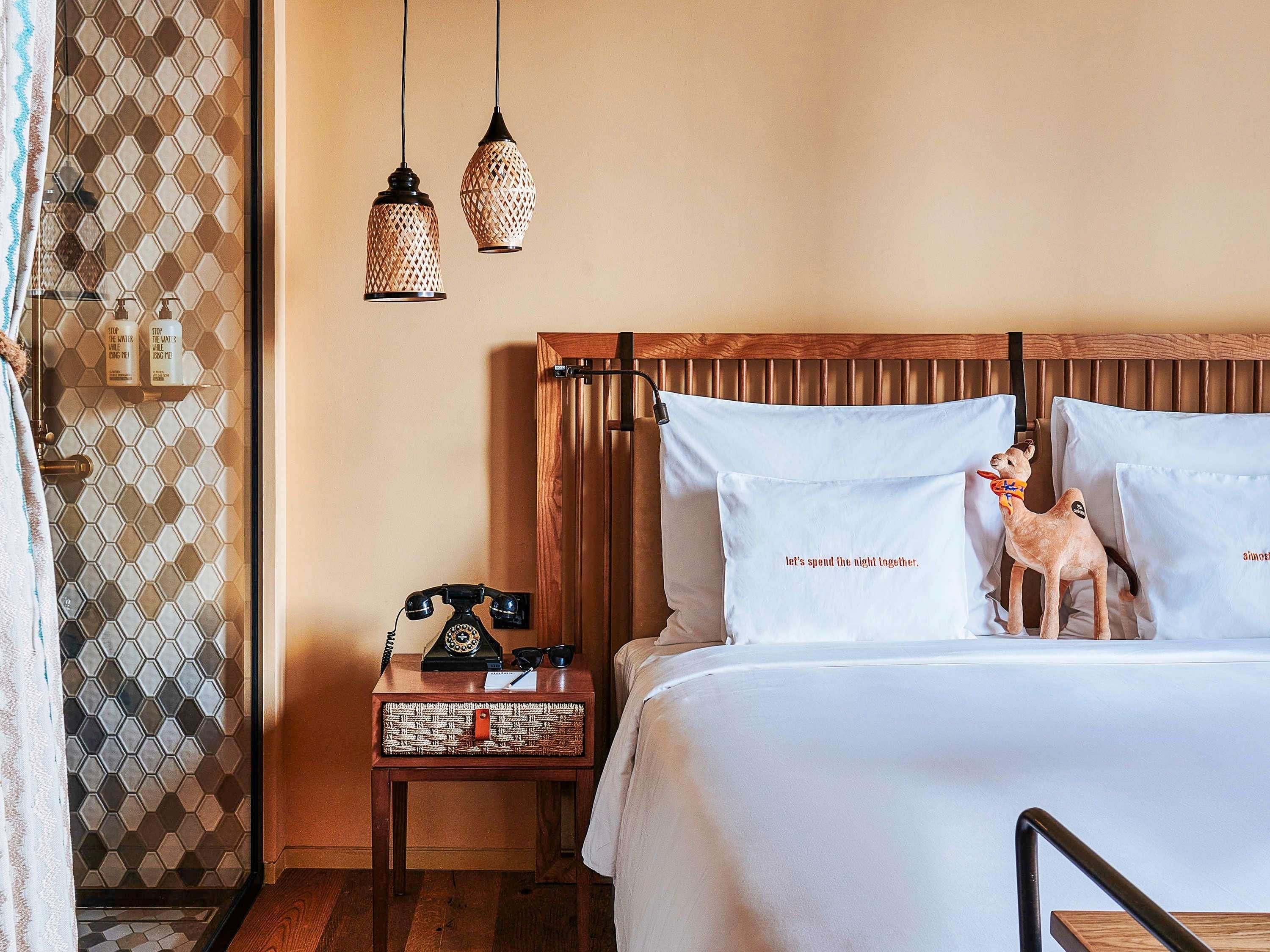 A neatly made bed with white bedding, a wooden headboard, and two decorative pillows with text. There is a small bedside table with a vintage telephone, a decorative item, and a bottle. Two hanging light fixtures with a woven design are visible.