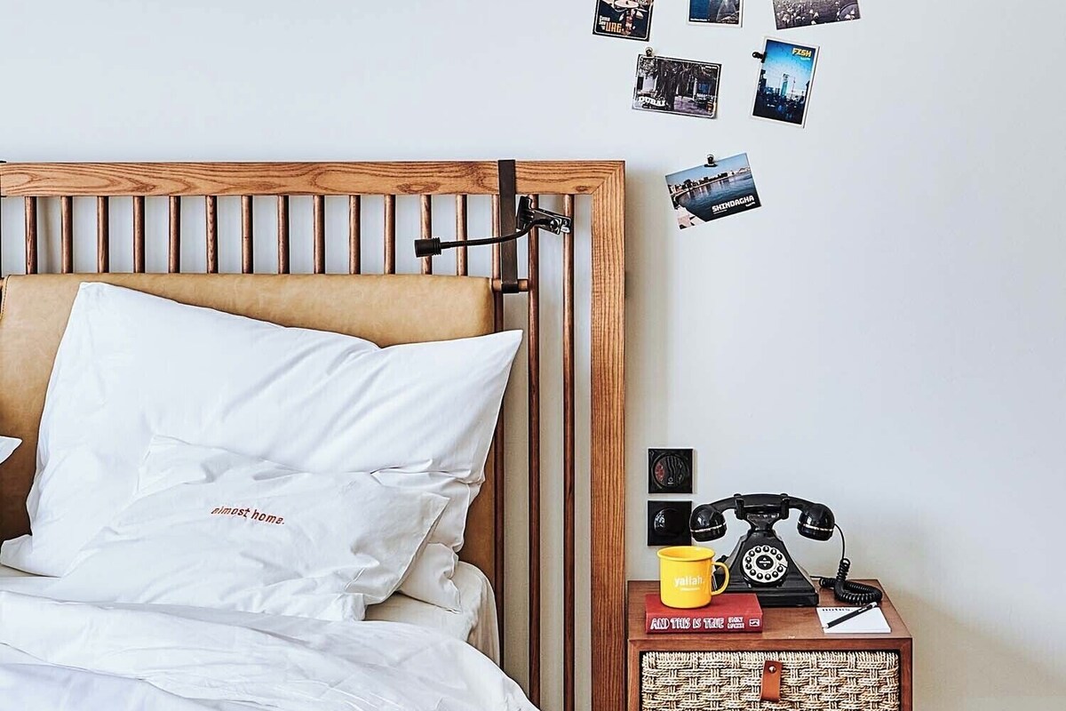 A bedroom with a bed, a wall with photos, a wooden nightstand with a phone and a yellow mug, and a wooden headboard.