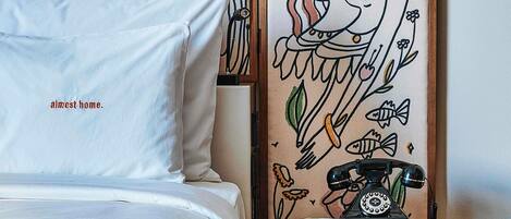 A hotel room with a bed, pillows, a bedside table with a book, a vintage telephone, and a tapestry with a floral design.