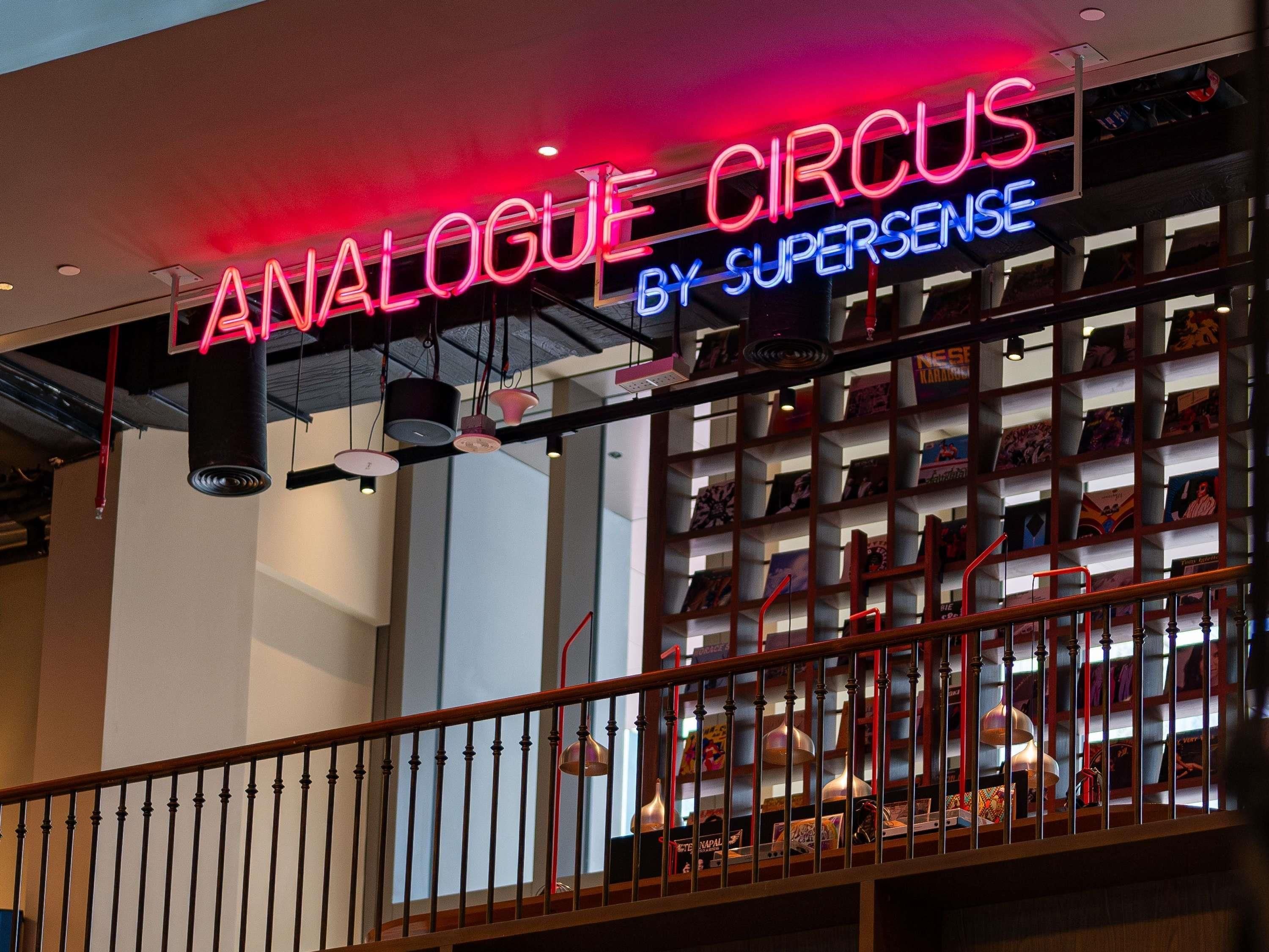 A neon sign reading 'ANALOGUE CIRCUS BY SUPERSENSE' above a bookshelf.