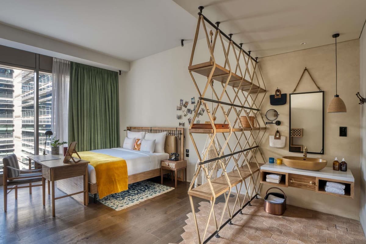 Moderni hotellihuone, jossa on puinen sänky, työpöytä, suuri ikkuna verhoilla, ainutlaatuinen geometrisen muotoinen hylly, pyöreä peili ja puinen pesuallas.