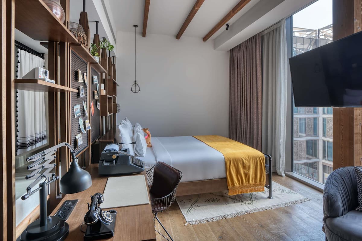 Moderni hotellihuone, jossa on sänky, työpöytä, tuoli ja suuri ikkuna.