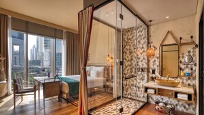 Habitación de hotel moderna con un ventanal grande, ducha envuelta en vidrio, piso de madera y vista a la ciudad.