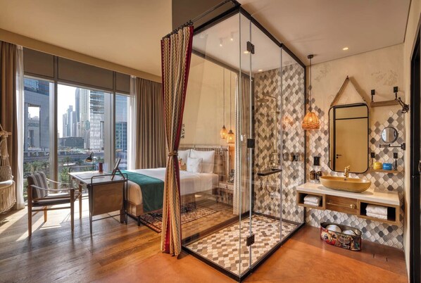 A modern hotel room with a large window, a glass-enclosed shower, a wooden floor, and a city view. - 25hours Hotel Dubai One Central (Dubai)