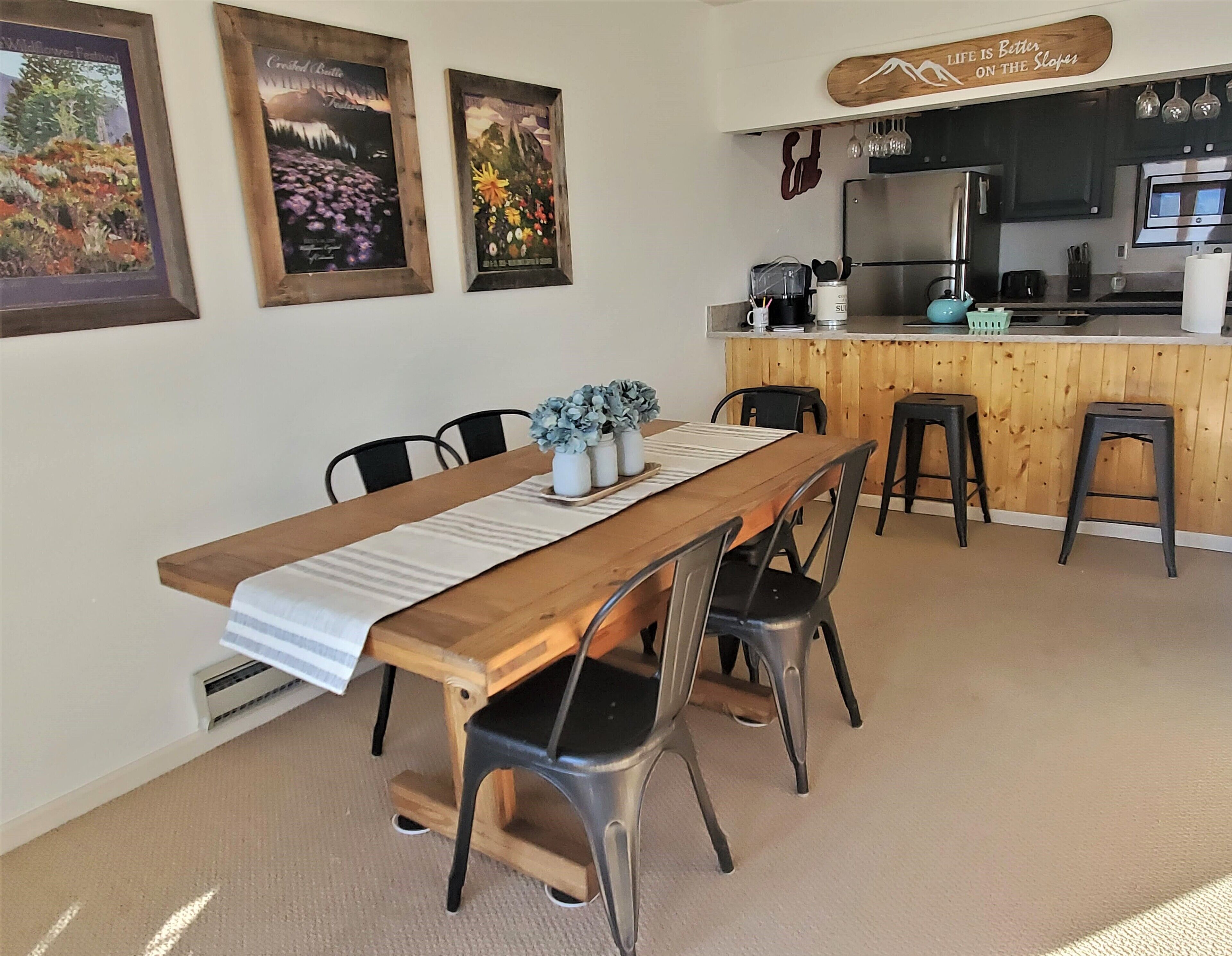 A dining area with a long wooden table, four chairs, and a bench. There are three framed pictures on the wall, a kitchen area with a refrigerator, stove, and sink, and a sign above the counter.