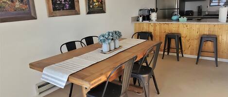 A dining area with a long wooden table, four chairs, and a bench. There are three framed pictures on the wall, a kitchen area with a refrigerator, stove, and sink, and a sign above the counter.