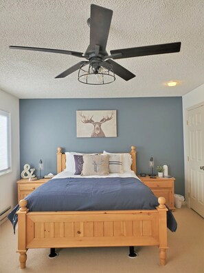 Quarto com cama de madeira, ventilador de teto, obra de arte na parede com um alce e mesa de cabeceira com abajur e relógio.