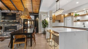 A modern kitchen with a central island, wooden beams, a stone fireplace, and a chandelier.