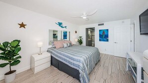 A bedroom with a bed, a nightstand, a potted plant, a ceiling fan, a door, and a wall-mounted TV. - Waterfront 1BR condo with balcony, pool, hot tub, and resort perks in St. Petersburg. (St Petersburg)