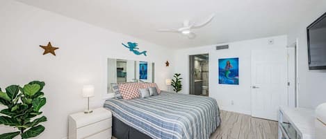 A bedroom with a bed, a nightstand, a potted plant, a ceiling fan, a door, and a wall-mounted TV.