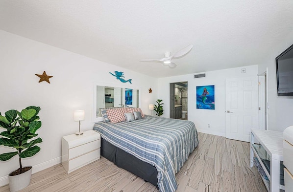 A bedroom with a bed, a nightstand, a potted plant, a ceiling fan, a door, and a wall-mounted TV.