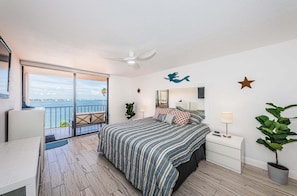 A bedroom with a bed, bedside tables, a nightstand, a ceiling fan, a large window with a view, and a potted plant.