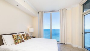A hotel room with a large bed, a nightstand with a lamp, a view of the sea through a sliding glass door, and a balcony. - Island and Sunset Views - P4-1008 (Pensacola Beach)