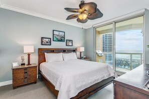A hotel room with a large bed, a wooden nightstand, a ceiling fan, and a large window with a city view. - P4-1106 Blue Bliss at Portofino with Gulf View! (Pensacola Beach)