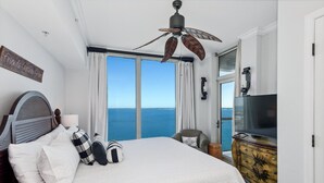A bedroom with a large bed, a ceiling fan, a television, and a view of the sea.