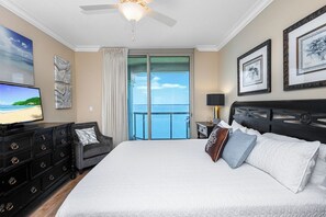 A hotel room with a large bed, a television, a dresser, and a balcony with a view of the ocean.