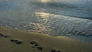 Ein Strand mit Fußspuren, die zum Wasser führen, einem klaren Himmel und der Sonne, die sich im Meer spiegelt.