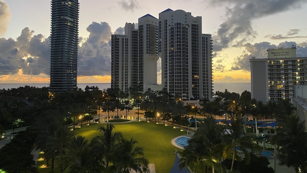 Ein Resort mit mehreren Hochhäusern, einem gut beleuchteten Poolbereich und Blick auf den Ozean bei Sonnenuntergang.