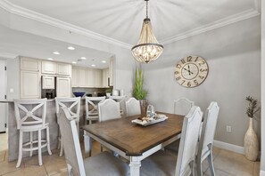 A dining area with a wooden table, white chairs, a chandelier, a clock, and a vase with plants.