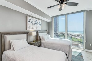Une chambre avec deux lits, un ventilateur de plafond, une table de chevet, une lampe et une grande fenêtre donnant sur la ville et la plage.