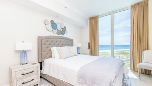 A bedroom with a large bed, two bedside tables, a nightstand, a chair, and a view of the ocean.