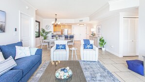 A living room with a blue sofa, striped armchairs, a coffee table with decorative items, a dining area with a chandelier, and a kitchen in the background.