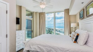 Un dormitorio con una cama grande, ventilador de techo, televisor y vistas a la ciudad a través de amplias ventanas.