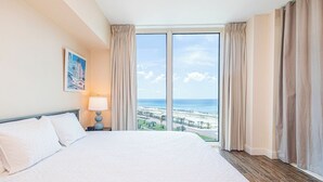 A hotel room with a large bed, a nightstand with a lamp, a painting on the wall, and a view of the ocean through a large window. - P5-0808 2B Gulf Front - Fabulous Gulf Front Views (Pensacola Beach)