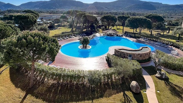 A circular swimming pool with a central island, surrounded by a stone deck and lounge chairs, set in a landscaped area with trees and a view of distant hills. - Apartment 85m2 luxury residence sea view 6 people 2 bedrooms (Porto-Vecchio)