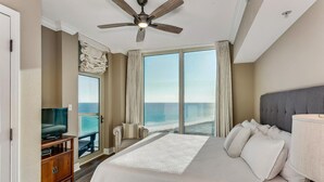 Habitación de hotel con una cama grande, ventilador de techo, televisor sobre un soporte de madera y vista al océano a través de ventanas amplias.