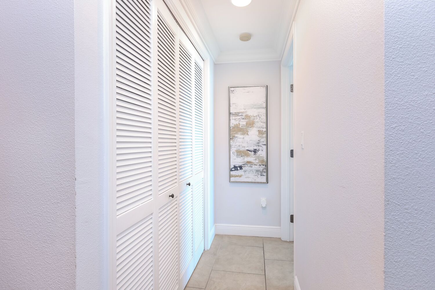 A narrow hallway with white walls, a tiled floor, and a white closet with louvered doors. A framed abstract painting hangs on the wall.