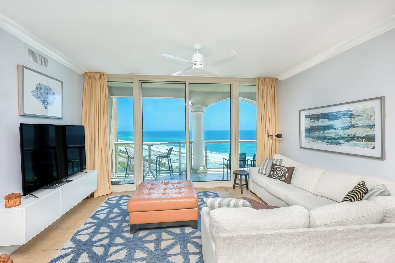 A living room with a TV, a sofa, an ottoman, and a view of the ocean through large glass doors.