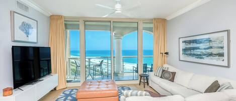 A living room with a TV, a sofa, an ottoman, and a view of the ocean through large glass doors.