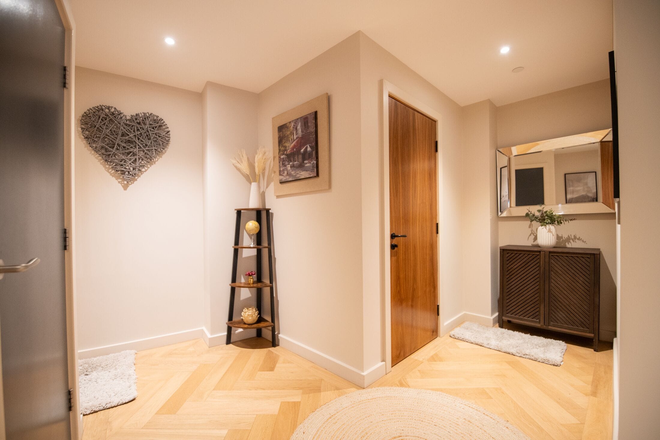 A hallway with a wooden door, a heart-shaped wall decoration, a framed picture, a shelf with decorative items, and a mirror above a cabinet.