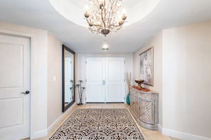 A hallway with a patterned rug, a chandelier, a door, a mirror, and a console table with a vase.