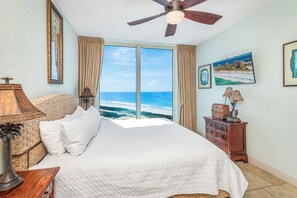 A bedroom with a bed, a ceiling fan, a nightstand, a lamp, a dresser, and a large window with a view of the ocean.