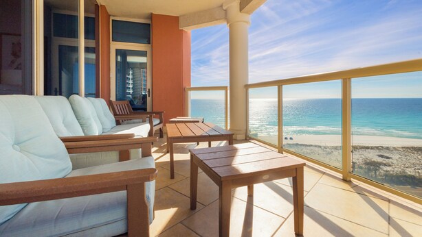 Un balcón con un sofá blanco, sillas de madera y una mesa, con vista a una playa, el océano y un cielo despejado.