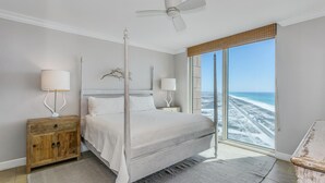 A bedroom with a bed, a nightstand, a lamp, a ceiling fan, and a large window with a view of the beach. - Gulf Front Views*Wraparound Balcony (Pensacola Beach)