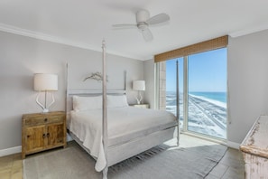 A bedroom with a bed, a nightstand, a lamp, a ceiling fan, and a large window with a view of the beach.