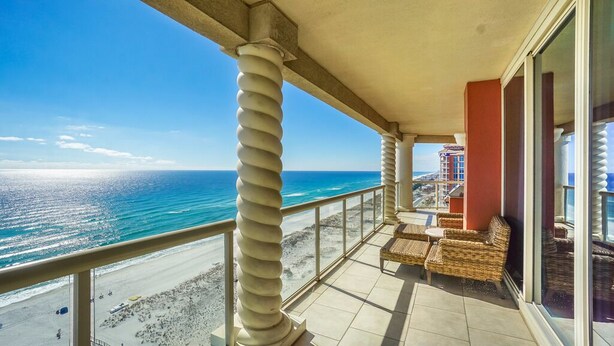 A balcony with a view of the ocean, featuring a seating area with wicker furniture and a glass railing. - P3-1901 Stunning 3B Gulf Front - Wrap Around Balcony! (Pensacola Beach)